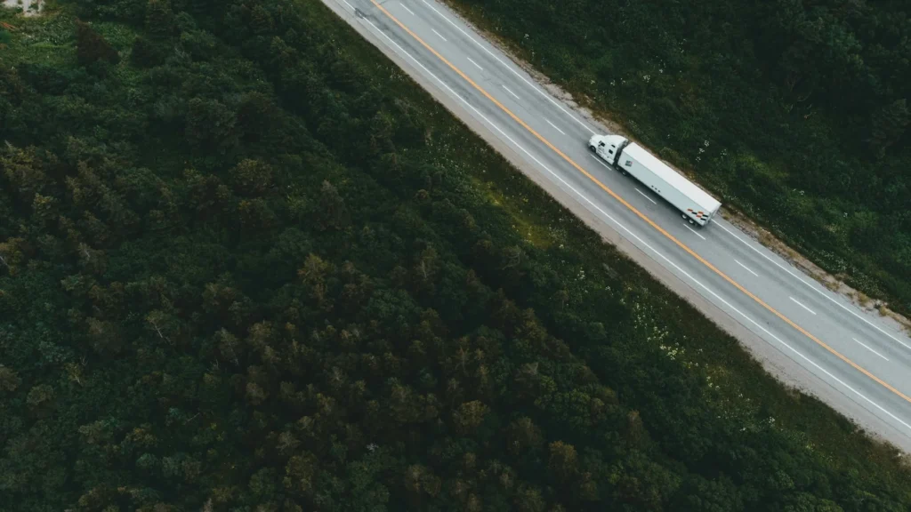 Белый грузовик едет по трассе между зеленых лесов