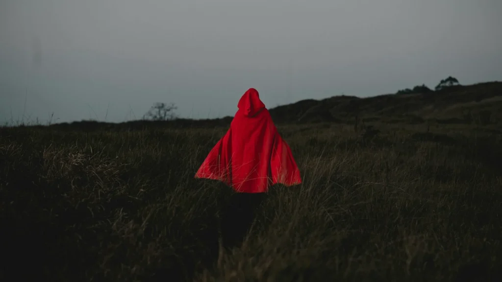 Человек в красном плаще с капюшоном идет по полю в сумерках
