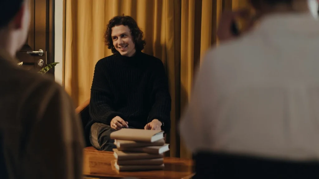 Молодой человек в черном выступает перед публикой, на столе книги