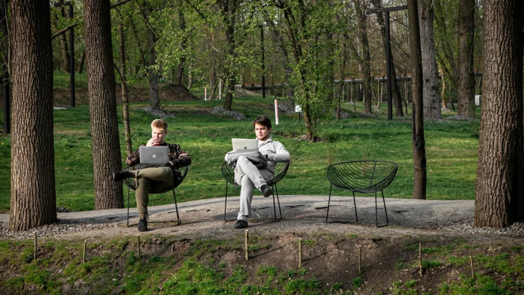 два молодых человека сидят в креслах в парке и работают за ноутбуками