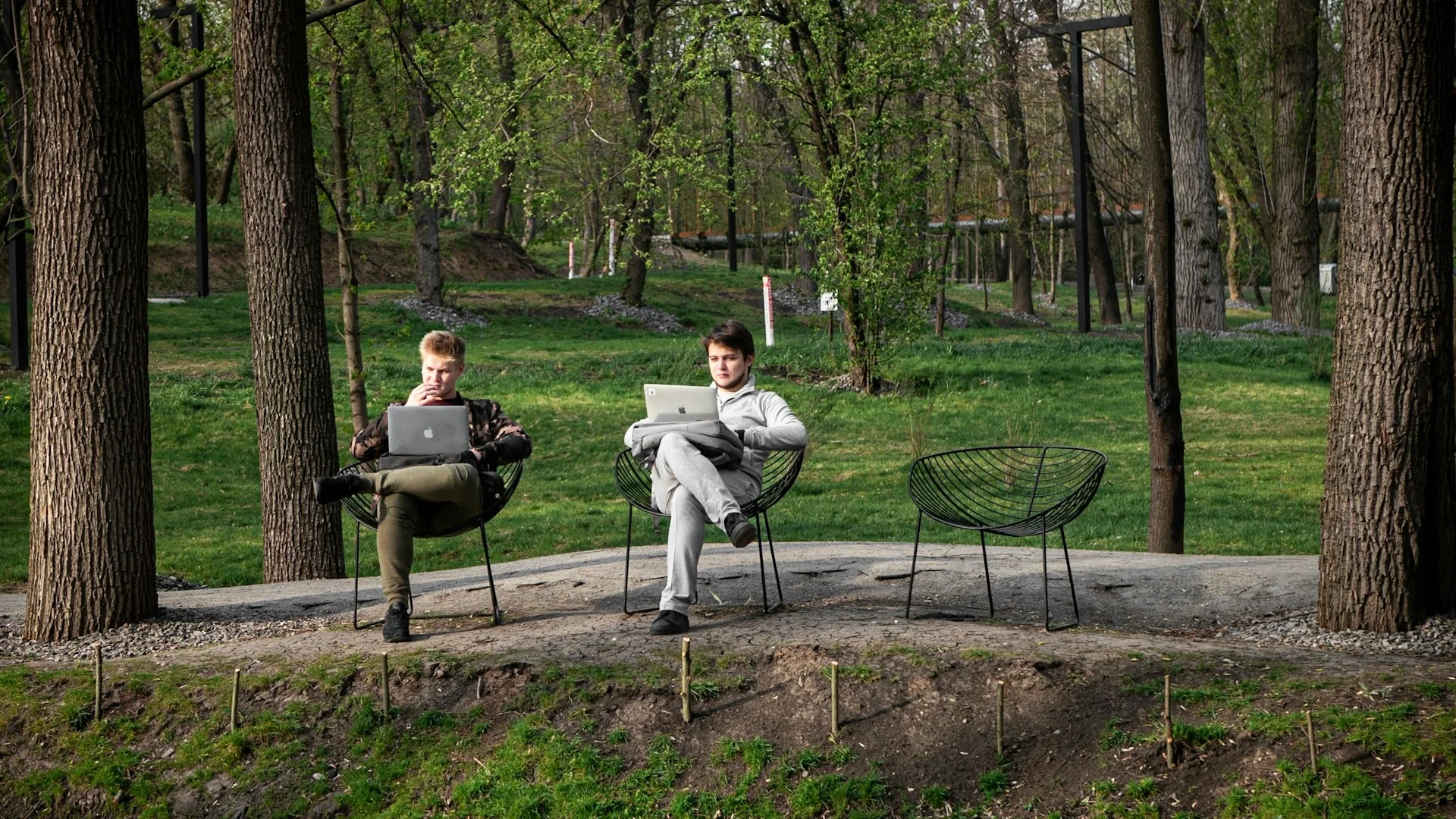 два молодых человека сидят в креслах в парке и работают за ноутбуками
