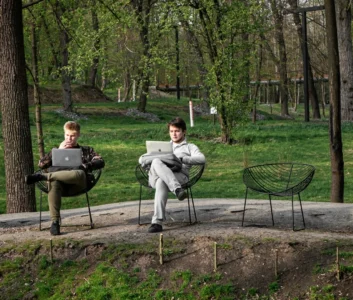 два молодых человека сидят в креслах в парке и работают за ноутбуками