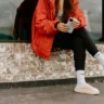 Днвушка в яркой куртке сидит на парапете с телефоном в руках
